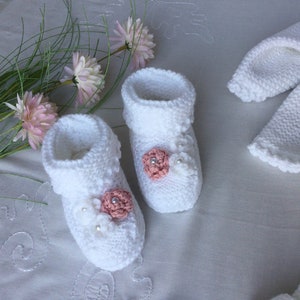 May include: White knitted baby booties with pink flower embellishments and white pearl accents. The booties are paired with a matching white knitted hat.