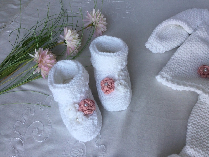 May include: White knitted baby booties with pink flower embellishments and white pearl accents. The booties are paired with a matching white knitted hat.