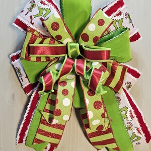 Peut inclure: Un nœud décoratif composé de plusieurs couches de ruban armé vert lime, rouge foncé et blanc. Le design comprend du tissu vert uni, des motifs à rayures rouges et vertes, et un ruban blanc avec un imprimé répétitif de personnage.