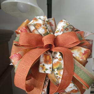 Puede incluir: Un lazo decorativo con múltiples capas de cinta en naranja, verde y blanco. Las cintas muestran patrones de calabazas, hojas de otoño y girasoles. Algunas secciones de cinta tienen una textura naranja sólida, mientras que otras son brillantes.