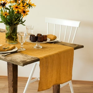 May include: A rustic wooden table with a mustard yellow linen table runner. The table is set with two place settings, including a plate, napkin, and a glass. There are also four small cakes on a cake stand.
