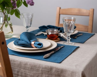 May include: A table setting with a blue linen napkin, a white plate, a blue placemat, and a glass of water. There is a small jar of honey on the table.