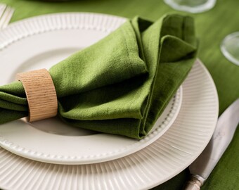 May include: A table setting featuring white plates with beaded and ribbed rims, a folded green linen napkin held by a wooden ring, and a silver knife. The table is covered with a green tablecloth.