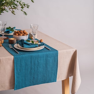 May include: A teal linen table runner on a beige linen tablecloth with a place setting, including a white plate, a blue napkin, a fork, and a glass. There is a small bowl of walnuts on the table.