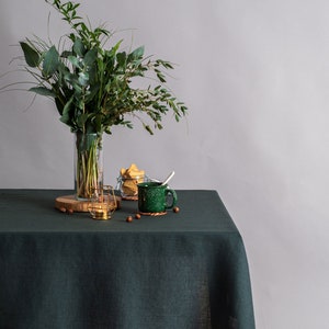 Könnte beinhalten: Ein Stillleben mit einer dunkelgrünen Leinentischdecke. Eine klare Glasvase enthält einen Strauß Grünzeug. Eine grüne Tasse, ein Glas mit Keksen und ein goldener Kerzenhalter stehen ebenfalls auf dem Tisch.