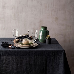May include: A black linen tablecloth with a place setting featuring a glass of water, a small jar with a sprig of rosemary, a plate, and a napkin. A green vase and a small cactus in a black pot are on the table.