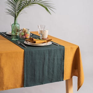 May include: A dining table set with a mustard yellow tablecloth and a dark teal runner. The table is set with plates, napkins, silverware, and glassware. A green palm frond is in a green glass bottle.