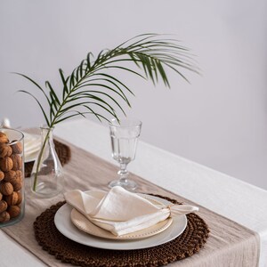 Op de afbeelding: Een witte linnen servet gevouwen op een wit bord met een bruine geweven placemat. Een glas water en een vaas met een palmblad staan op tafel.