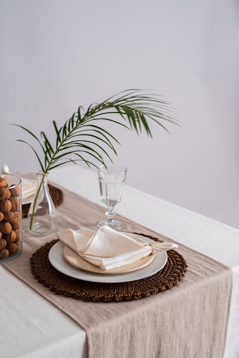 Op de afbeelding: Een witte linnen servet gevouwen op een wit bord met een bruine geweven placemat. Een glas water en een vaas met een palmblad staan op tafel.