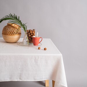 Op de afbeelding: Een wit tafelkleed bedekt een houten tafel met een geweven mand, een glazen pot gevuld met bruine noten, een heldere glazen vaas en een rode keramische mok. De tafel staat tegen een grijze achtergrond.