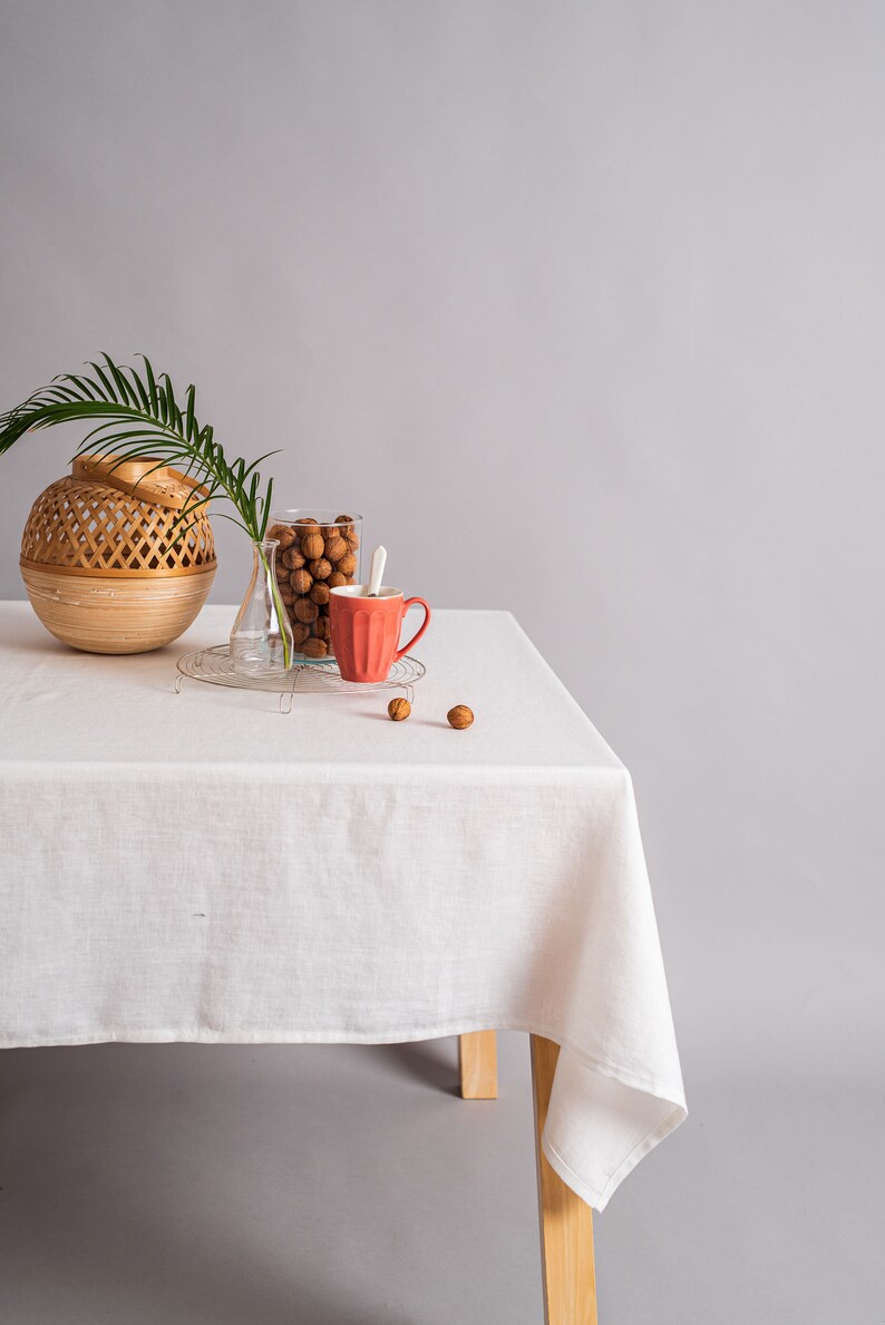 Op de afbeelding: Een wit tafelkleed bedekt een houten tafel met een geweven mand, een glazen pot gevuld met bruine noten, een heldere glazen vaas en een rode keramische mok. De tafel staat tegen een grijze achtergrond.
