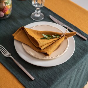 Pu&ograve; includere: Una tavola apparecchiata con un tovagliolo di lino color senape su un piatto bianco, decorato con un rametto di verde. La tavola &egrave; coperta da una tovaglia di lino verde scuro e senape. Sono visibili anche un bicchiere, posate e un barattolo.