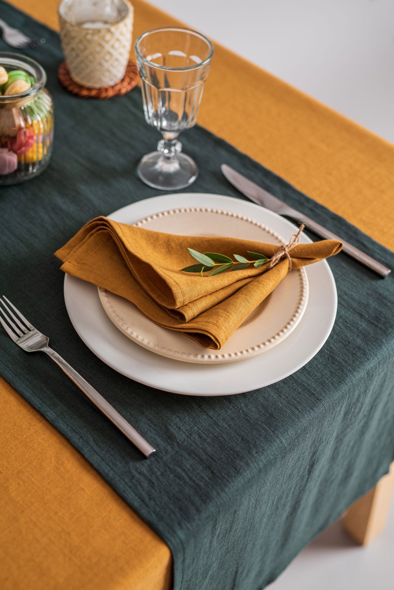 Pu&ograve; includere: Una tavola apparecchiata con un tovagliolo di lino color senape su un piatto bianco, decorato con un rametto di verde. La tavola &egrave; coperta da una tovaglia di lino verde scuro e senape. Sono visibili anche un bicchiere, posate e un barattolo.