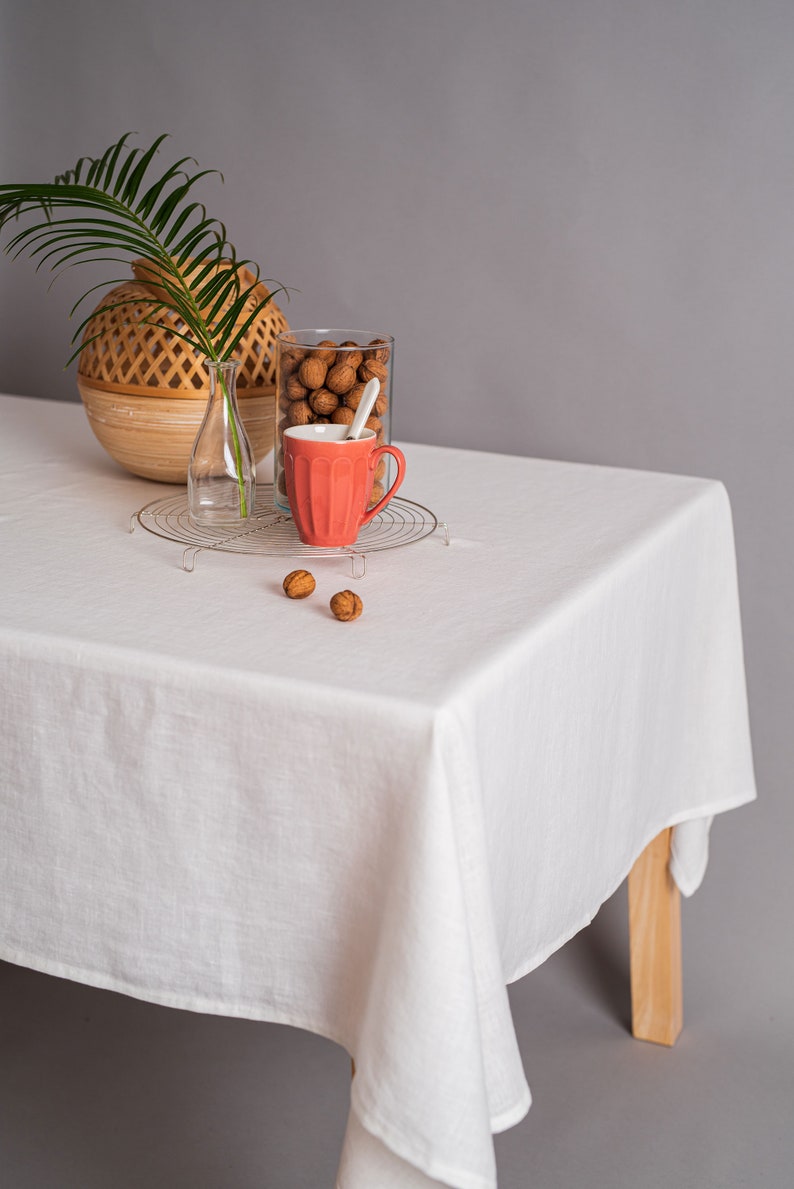 Op de afbeelding: Een wit tafelkleed bedekt een houten tafel met een geweven mand, een glazen vaas met een groene plant, een glazen pot gevuld met walnoten en een rode keramische mok op een draadrek. Er liggen drie walnoten op de tafel.