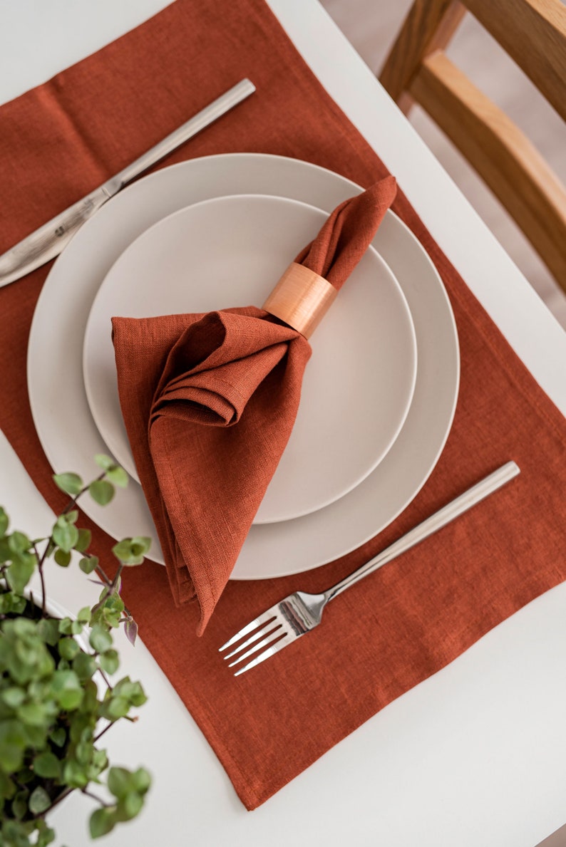 May include: A rust-colored linen placemat with a white plate and a folded linen napkin with a copper napkin ring. A silver fork is placed on the placemat.