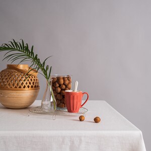 Op de afbeelding: Een wit tafelkleed met een rode mok, een glazen pot gevuld met walnoten, een glazen vaas en een geweven mand op een tafel. Er liggen ook een paar walnoten verspreid over de tafel.