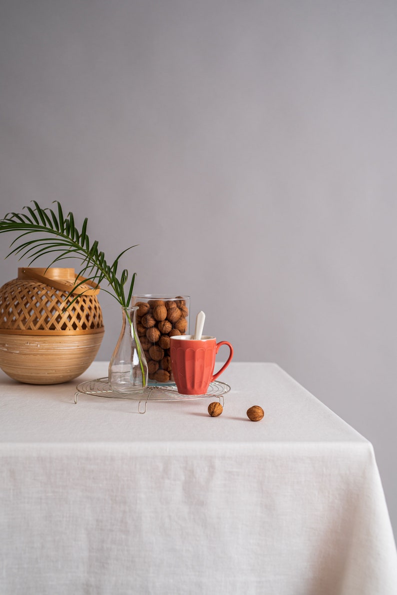 Op de afbeelding: Een wit tafelkleed met een rode mok, een glazen pot gevuld met walnoten, een glazen vaas en een geweven mand op een tafel. Er liggen ook een paar walnoten verspreid over de tafel.