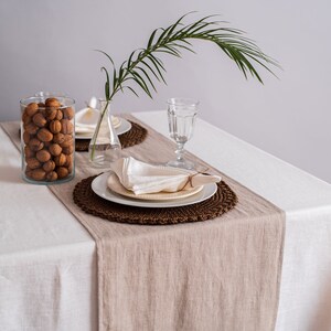 Op de afbeelding: Een wit tafelkleed met een bruine linnen loper. Een wit bord met een witte servet ligt op een bruin geweven placemat. Een glas water en een vaas met een palmblad staan op de tafel.