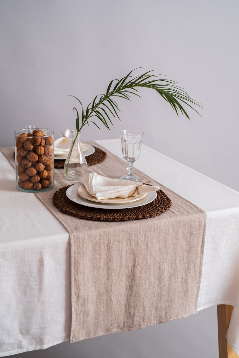 Op de afbeelding: Een wit tafelkleed met een bruine linnen loper. Een wit bord met een witte servet ligt op een bruin geweven placemat. Een glas water en een vaas met een palmblad staan op de tafel.