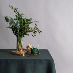 Pu&ograve; includere: Una natura morta con una tovaglia di lino verde scuro. Un vaso di vetro trasparente contiene un bouquet di foglie verdi. Una tazza verde con un cucchiaio, un piccolo barattolo di vetro con biscotti e un portacandele dorato sono anche sul tavolo.