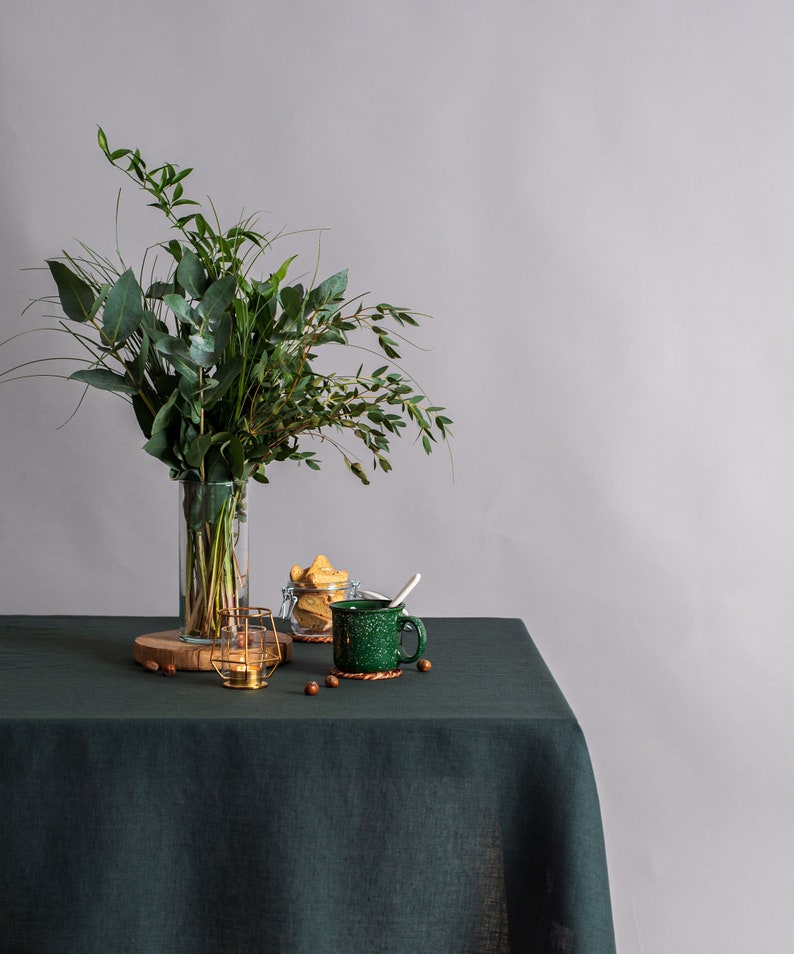 Pu&ograve; includere: Una natura morta con una tovaglia di lino verde scuro. Un vaso di vetro trasparente contiene un bouquet di foglie verdi. Una tazza verde con un cucchiaio, un piccolo barattolo di vetro con biscotti e un portacandele dorato sono anche sul tavolo.