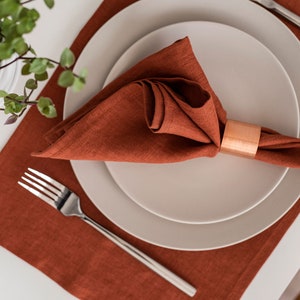 May include: A rust-colored linen napkin is folded and placed on a white plate with a copper napkin ring. The plate is on a rust-colored placemat.