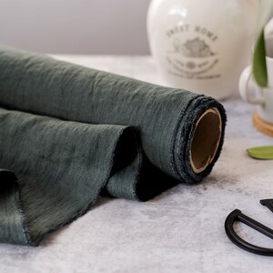 Pu&ograve; includere: Un rotolo di tessuto verde scuro con un bordo grezzo. Il tessuto &egrave; strutturato e sembra essere lino. Un paio di forbici nere sono in primo piano. Un boccale e una tazza di ceramica bianchi sono sullo sfondo.