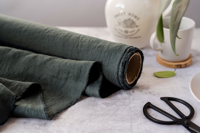 Pu&ograve; includere: Un rotolo di tessuto verde scuro con un bordo grezzo. Il tessuto &egrave; strutturato e sembra essere lino. Un paio di forbici nere sono in primo piano. Un boccale e una tazza di ceramica bianchi sono sullo sfondo.