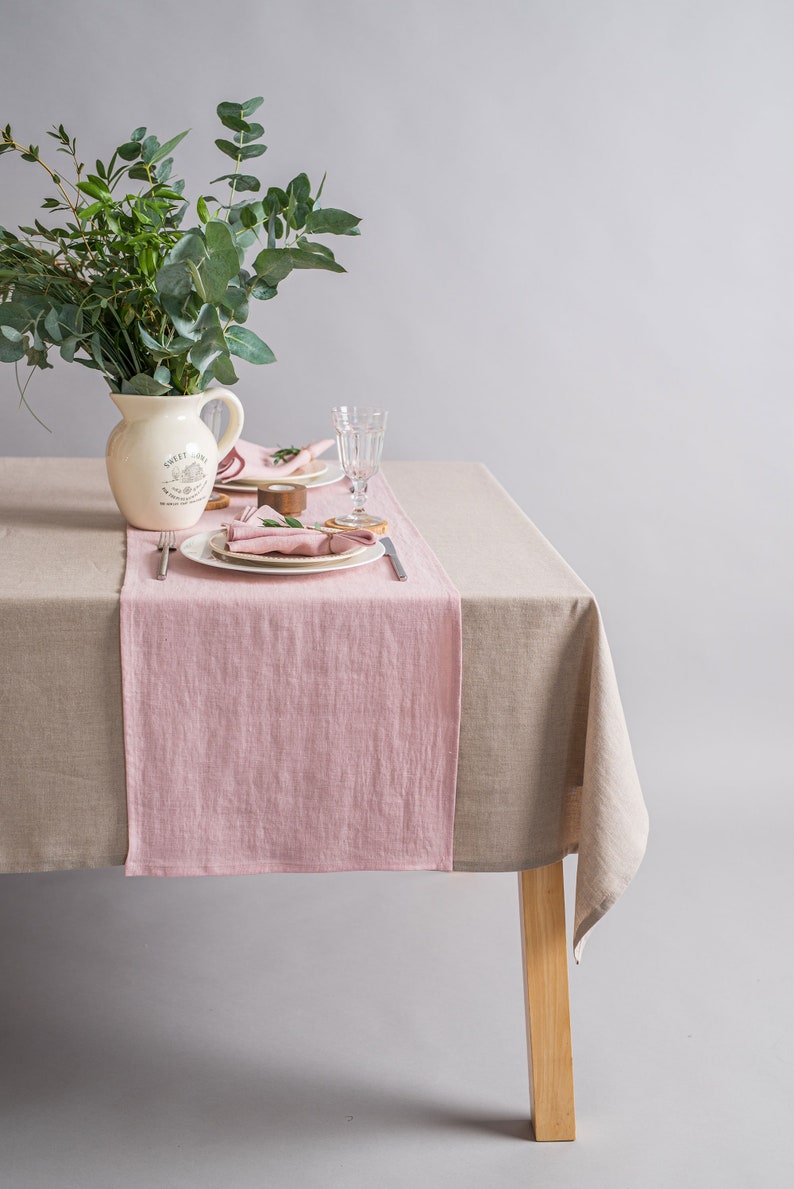 May include: A light pink linen table runner on a beige linen tablecloth. A white pitcher with the text "Sweet Home" is on the table. There is a glass of water and a plate with a pink napkin on the table.