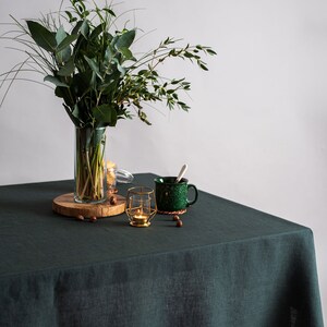 Pu&ograve; includere: Una tovaglia di lino verde scuro copre un tavolo di legno. Un vaso trasparente con fogliame verde &egrave; posto su una fetta di legno. Una tazza verde maculata, un portacandele dorato e nocciole sono sul tavolo.