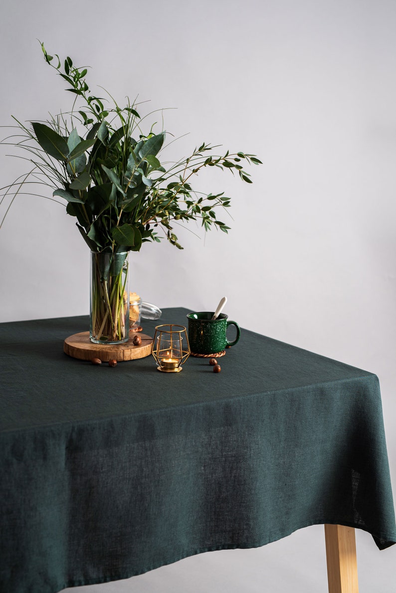 Pu&ograve; includere: Una tovaglia di lino verde scuro copre un tavolo di legno. Un vaso trasparente con fogliame verde &egrave; posto su una fetta di legno. Una tazza verde maculata, un portacandele dorato e nocciole sono sul tavolo.