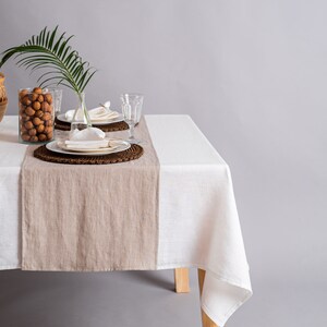 Op de afbeelding: Een wit linnen tafelkleed met een beige linnen loper op een houten tafel. De tafel is gedekt met twee couverts, elk met een wit bord, een witte servet en een helder glas. Een vaas met een groen palmblad en een kom met walnoten staan op de tafel.