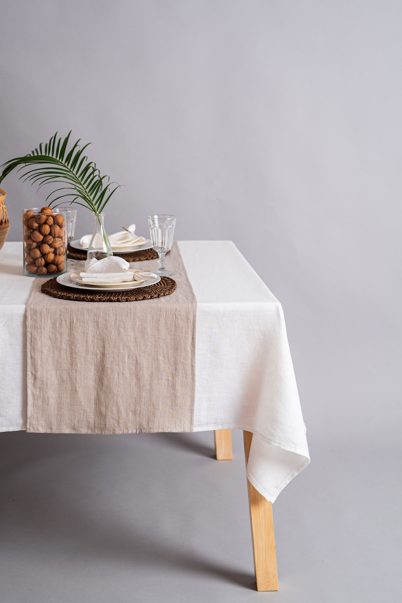 Op de afbeelding: Een wit linnen tafelkleed met een beige linnen loper op een houten tafel. De tafel is gedekt met twee couverts, elk met een wit bord, een witte servet en een helder glas. Een vaas met een groen palmblad en een kom met walnoten staan op de tafel.