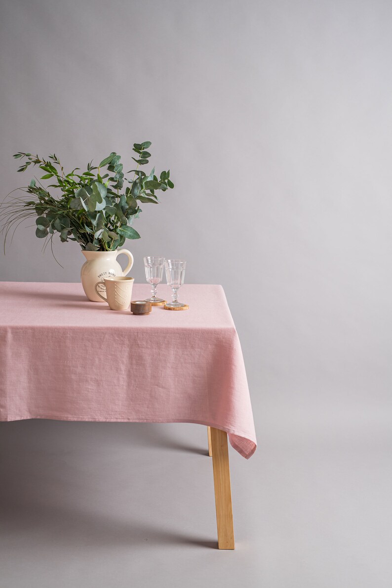 May include: A pink linen tablecloth on a wooden table with a white pitcher, two clear glasses, and a small wooden candle holder. The pitcher has the text "Keep it Real" on it. The table is set for a simple, elegant meal.
