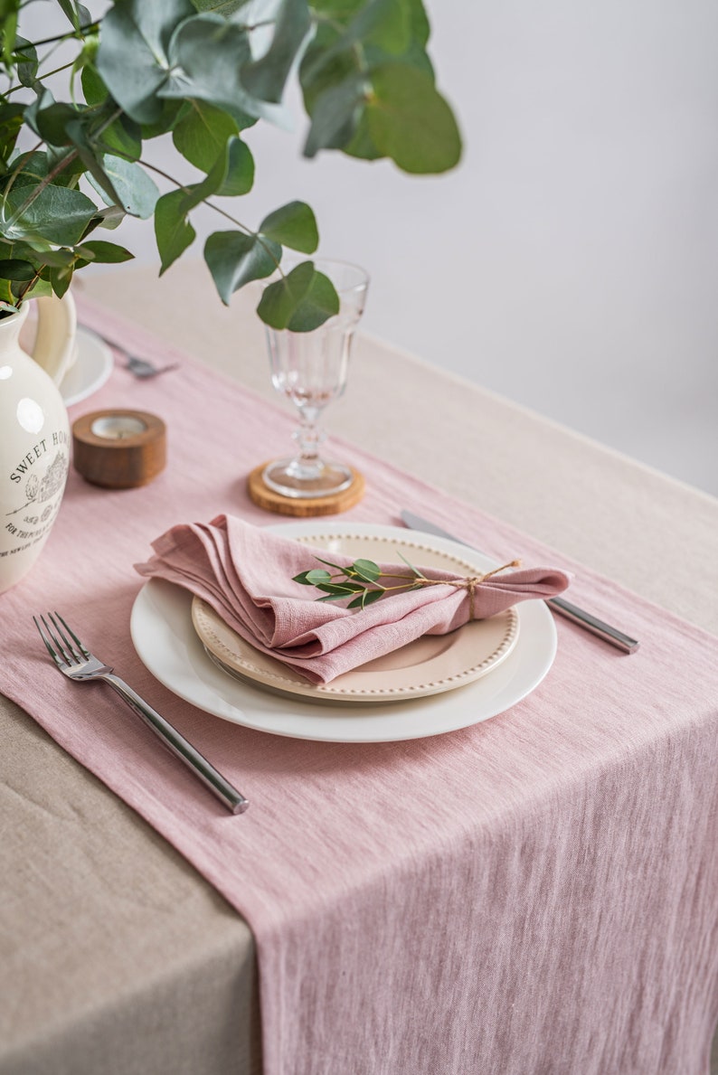May include: A pink linen table runner with a white plate and a pink linen napkin. A sprig of greenery is placed on the napkin. The table runner is on top of a beige tablecloth.
