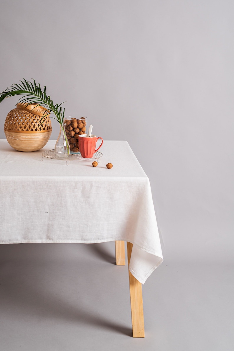 Op de afbeelding: Een wit linnen tafelkleed gedrapeerd over een houten tafel met een geweven mand, een glazen vaas, een glazen pot gevuld met bruine noten en een rode keramische mok. De tafel staat op een grijze achtergrond.
