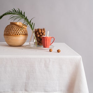 Op de afbeelding: Een wit tafelkleed bedekt een houten tafel met een geweven mand, een glazen pot gevuld met walnoten en een rode mok. De tafel staat tegen een grijze achtergrond.