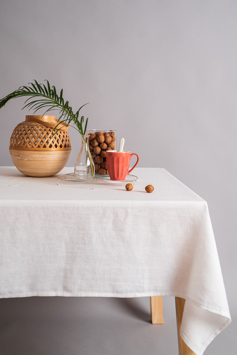 Op de afbeelding: Een wit tafelkleed bedekt een houten tafel met een geweven mand, een glazen pot gevuld met walnoten en een rode mok. De tafel staat tegen een grijze achtergrond.
