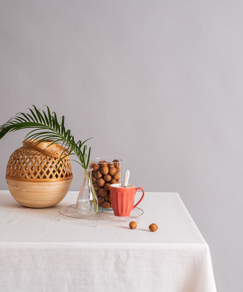 Op de afbeelding: Een wit tafelkleed met een geweven rieten mand, een glazen pot gevuld met walnoten, een heldere glazen vaas, een rode keramische mok en een paar walnoten die op de tafel zijn verspreid.
