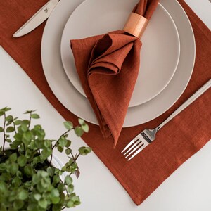 May include: A rust-colored linen placemat with a white plate and a folded linen napkin with a copper napkin ring. A silver fork and knife are on the placemat.