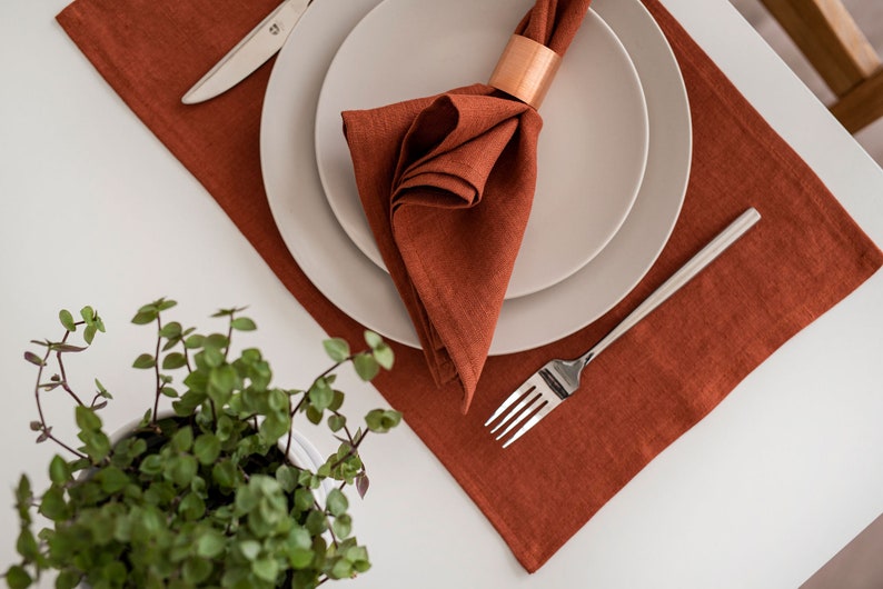 May include: A rust-colored linen placemat with a white plate and a folded linen napkin with a copper napkin ring. A silver fork and knife are on the placemat.