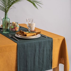Pu&ograve; includere: Una tavola apparecchiata con una tovaglia giallo senape e un runner verde scuro. Piatti bianchi con tovaglioli senape piegati, posate e bicchieri sono disposti. Una bottiglia di vetro verde con una foglia di palma &egrave; sullo sfondo.