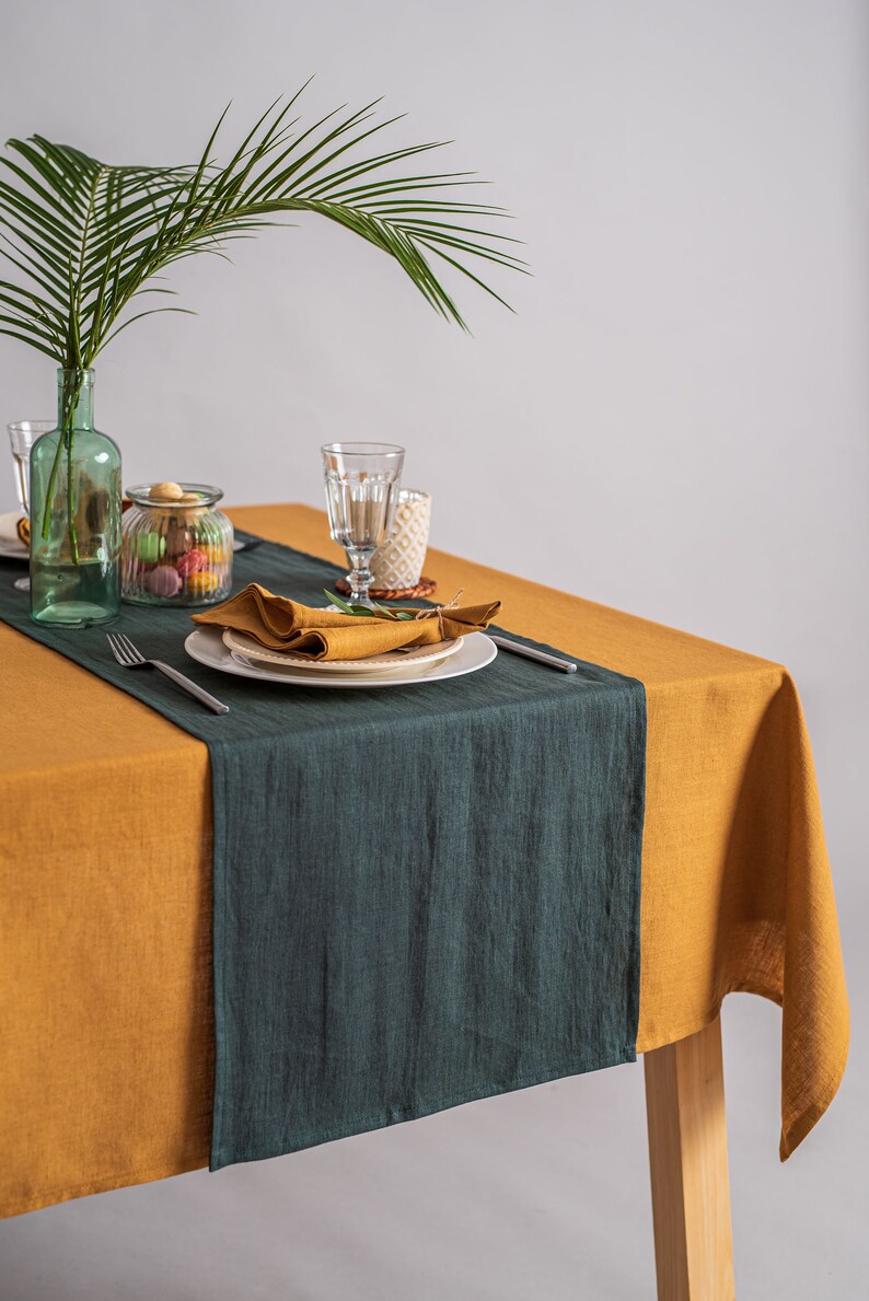 Pu&ograve; includere: Una tavola apparecchiata con una tovaglia giallo senape e un runner verde scuro. Piatti bianchi con tovaglioli senape piegati, posate e bicchieri sono disposti. Una bottiglia di vetro verde con una foglia di palma &egrave; sullo sfondo.