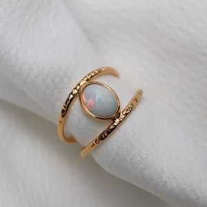 Peut inclure: Une bague en or avec une pierre d'opale ovale. La bague a un anneau texturé et un design à double anneau. L'opale présente un jeu de couleurs, sur fond blanc.