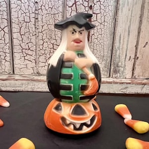 Peut inclure: Une figurine de sorcière en plastique moulé avec de longs cheveux blancs, un chapeau noir et un corsage rayé vert et noir, tenant un balai. La sorcière est placée sur une base de citrouille d'Halloween orange avec un visage sculpté.