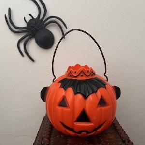 May include: An orange plastic Halloween pumpkin bucket with a black bat design and handle. A black plastic spider is above the bucket. The bucket has a smiling face and is a Halloween decoration.
