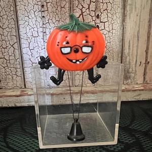 May include: A vintage-style orange pumpkin figure with a green stem, black limbs, and white-rimmed glasses. The pumpkin has a smiling face with visible teeth and is suspended above a clear acrylic cube by a thin black wire attached to a small black bell.