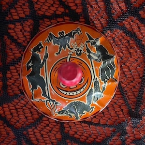 Puede incluir: Una peonza de juguete vintage con temática de Halloween con un botón rojo. La parte superior naranja presenta siluetas negras de una bruja, un murciélago, una araña y otras figuras espeluznantes. El fondo es un patrón de telaraña rojo y negro.