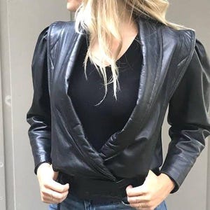 Op de afbeelding: Zwarte leren jas met een wikkelvoorkant en opgevulde schouders. De jas heeft lange mouwen en een ceintuur in de taille. Het model draagt een zwarte top en een blauwe jeans.
