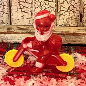 Może przedstawiać: Plastikowa figurka Świętego Mikołaja w kolorze czerwonym, jadąca na czerwonym trójkołowym rowerze z żółtymi kołami. Figurka ma białą brodę i czerwony kapelusz.